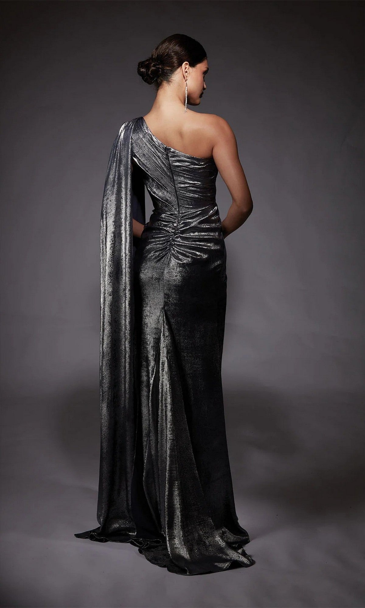 A woman faces away, wearing the Alyce 27753 long formal dress with metallic chiffon, a one-shoulder design, and a draped train, set against a plain dark background.